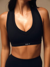A close-up of a woman wearing a black athletic sports bra with a wide scoop neckline and the brand name 'SET' printed in white on the underband, paired with black athletic bottoms against a warm beige background. 