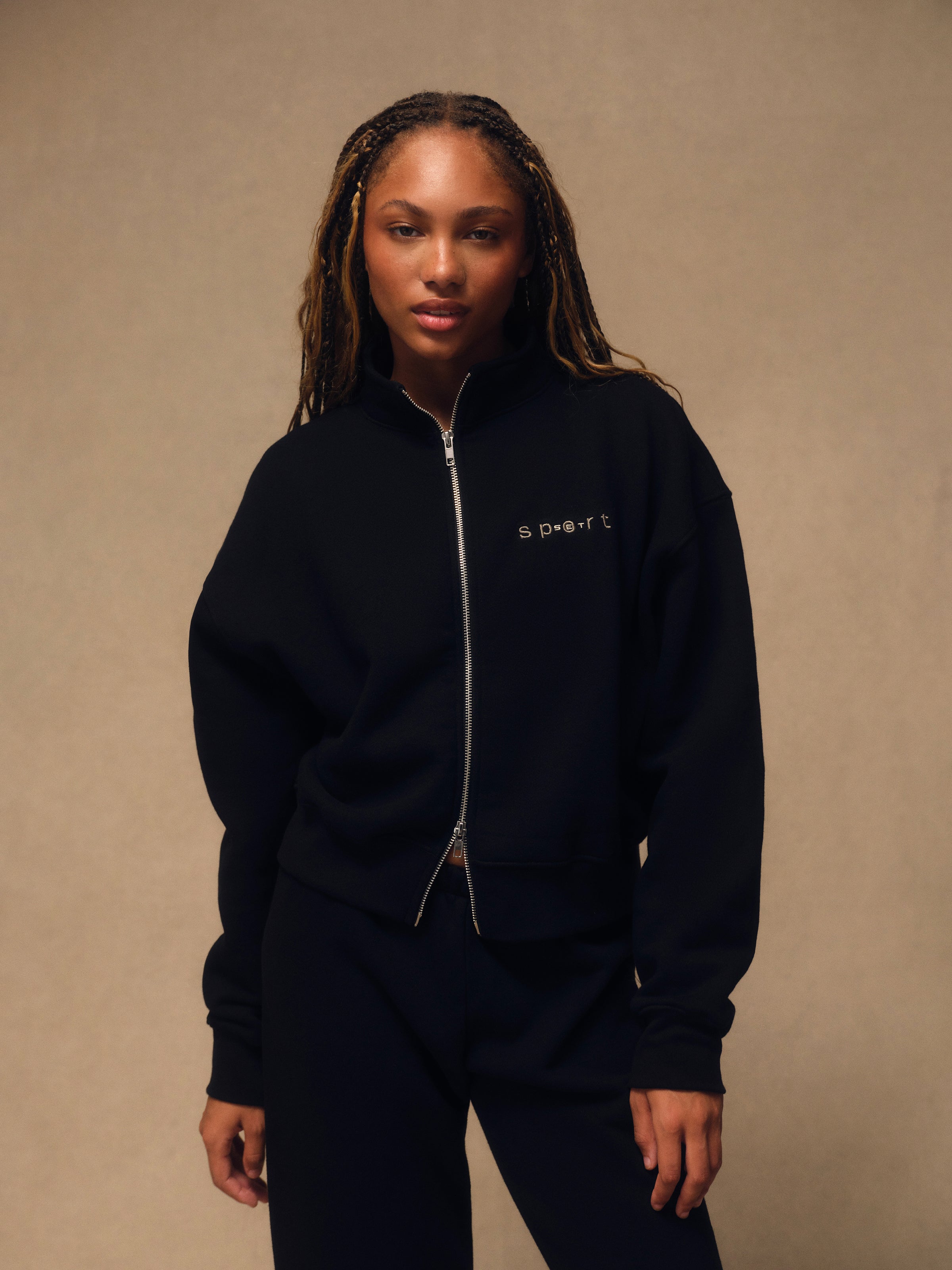 A woman wearing a black zip-up sweatshirt and matching black sweatpants, standing against a warm beige studio background. The sweatshirt has a small white embroidered logo on the left chest. 
