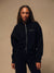 A woman wearing a black zip-up sweatshirt and matching black sweatpants, standing against a warm beige studio background. The sweatshirt has a small white embroidered logo on the left chest. <model-info>Lena wears a size XS and is 5’7”, 24½” waist, 31" bust.</model-info>
