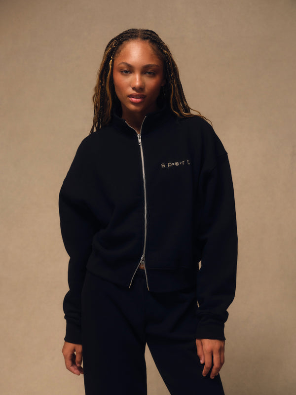 A woman wearing a black zip-up sweatshirt and matching black sweatpants, standing against a warm beige studio background. The sweatshirt has a small white embroidered logo on the left chest. 