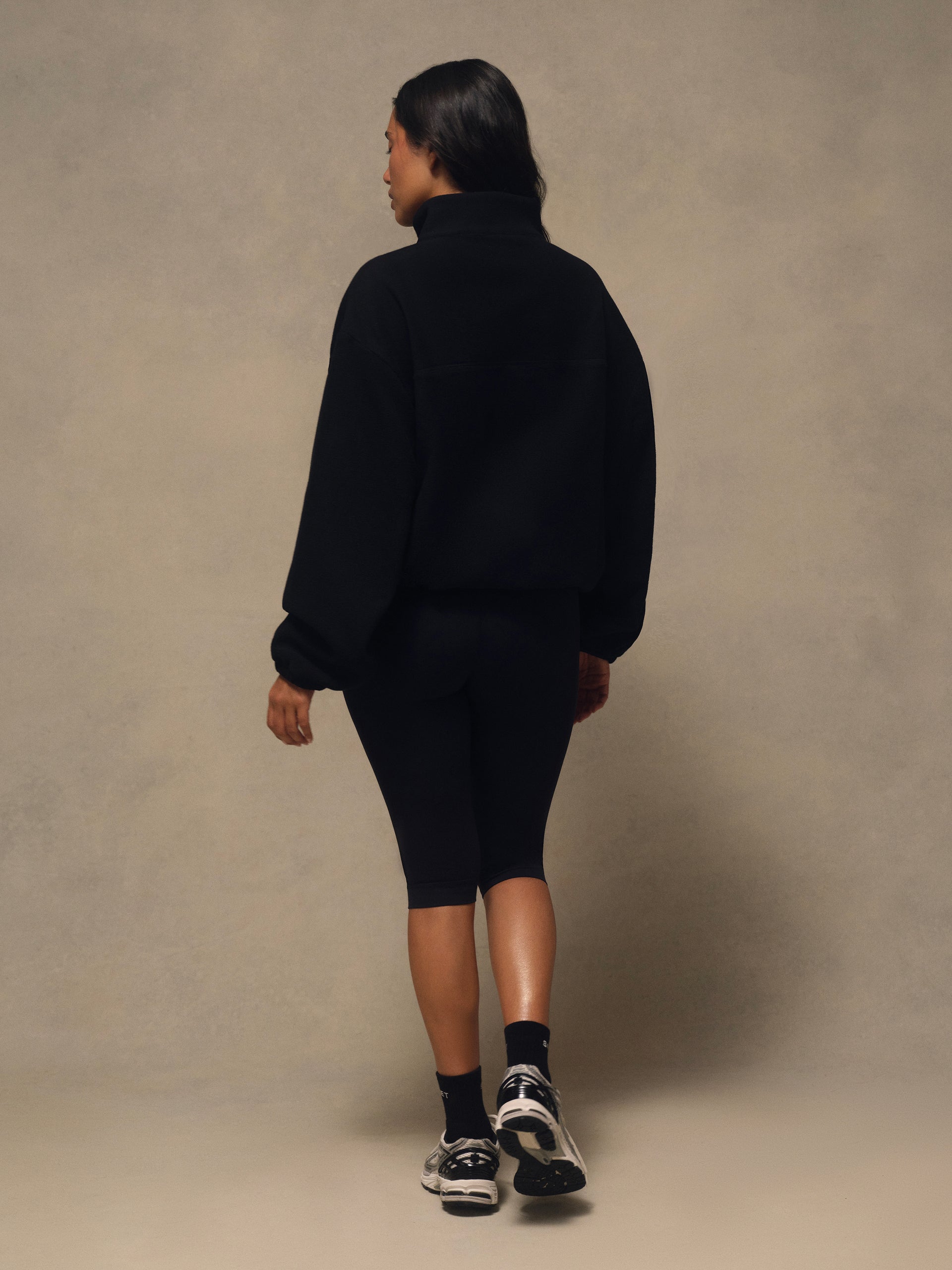 A model shown from behind wearing a black oversized mock-neck sweatshirt and matching black bike shorts, styled with black socks and black and white sneakers, against a textured beige studio backdrop.