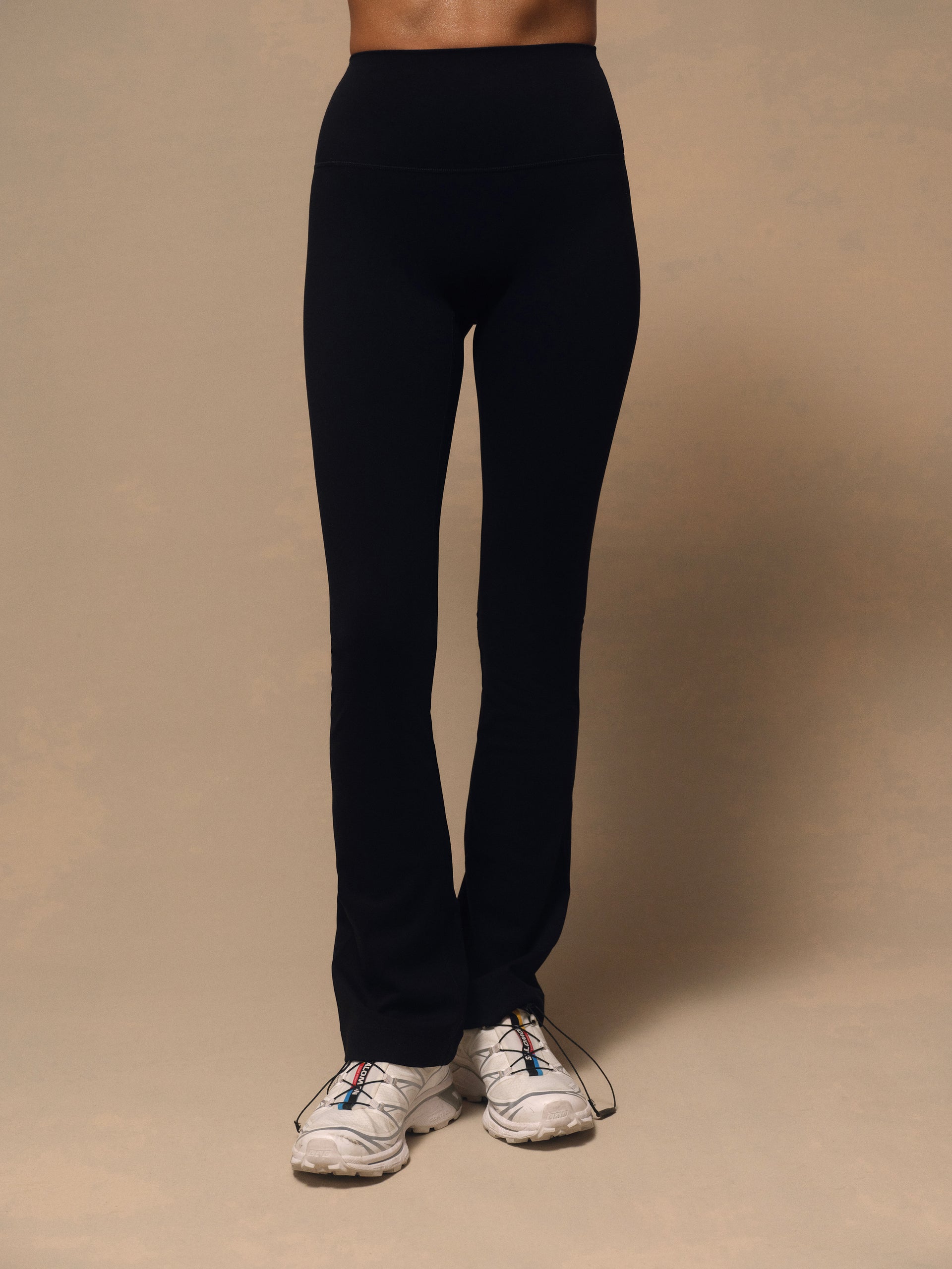 Black high-waisted athletic leggings worn by a model, paired with white trail running shoes, against a solid beige studio background.