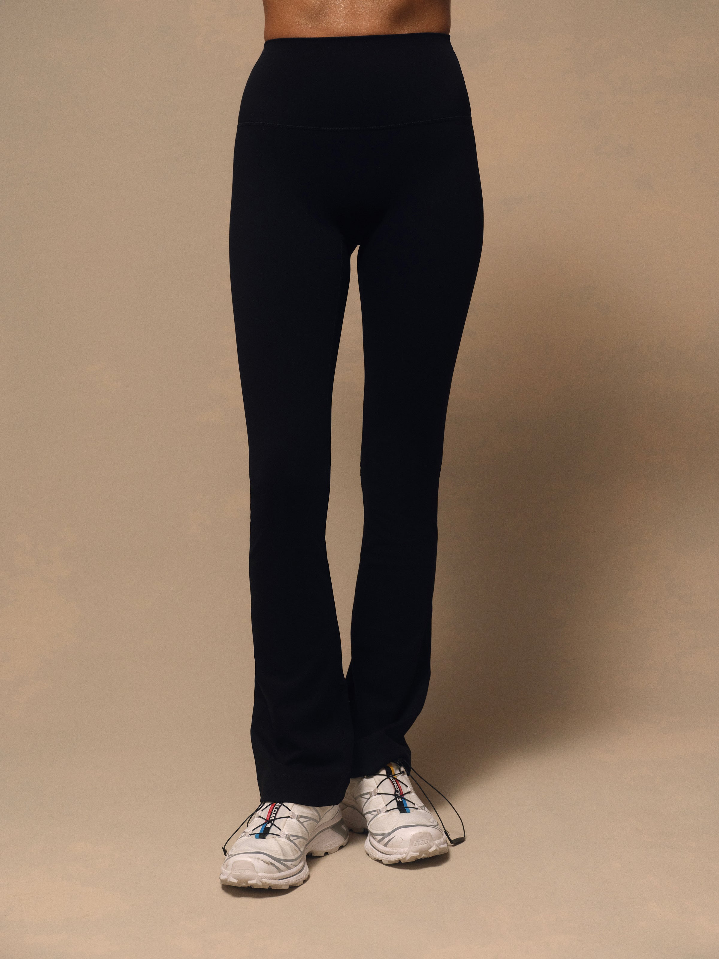 Black high-waisted athletic leggings worn by a model, paired with white trail running shoes, against a solid beige studio background.