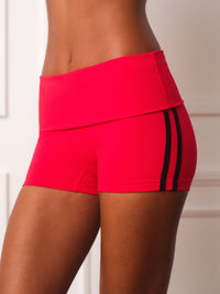 A close-up of a model wearing bright red athletic shorts with a wide waistband and black side stripes, shown against a light neutral indoor background. 