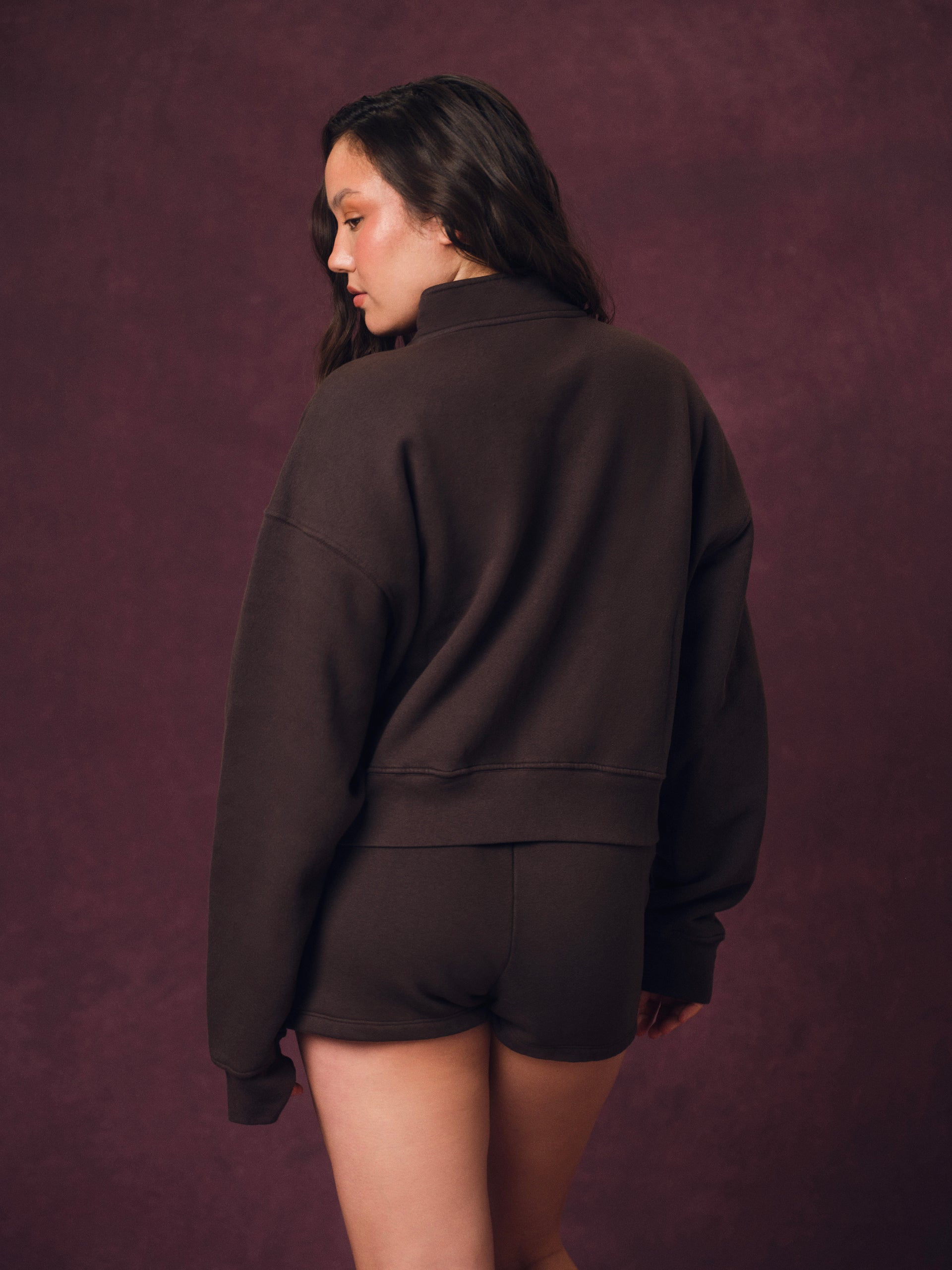 A woman wearing a dark brown cropped sweatshirt and matching shorts, photographed from the back against a textured dark plum studio backdrop.