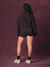 A model viewed from behind wearing a dark brown cropped sweatshirt and matching shorts, standing against a textured dark purple studio backdrop.