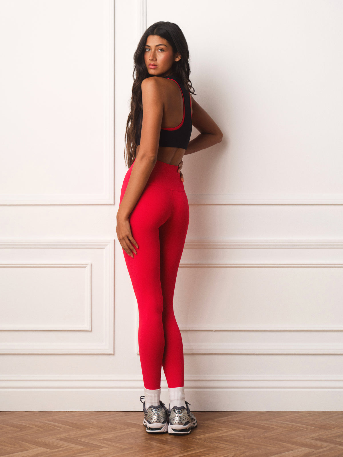 A woman stands in profile against a white paneled wall wearing bright red high-waisted leggings, a black sports bra with red trim, white socks, and gray athletic sneakers.