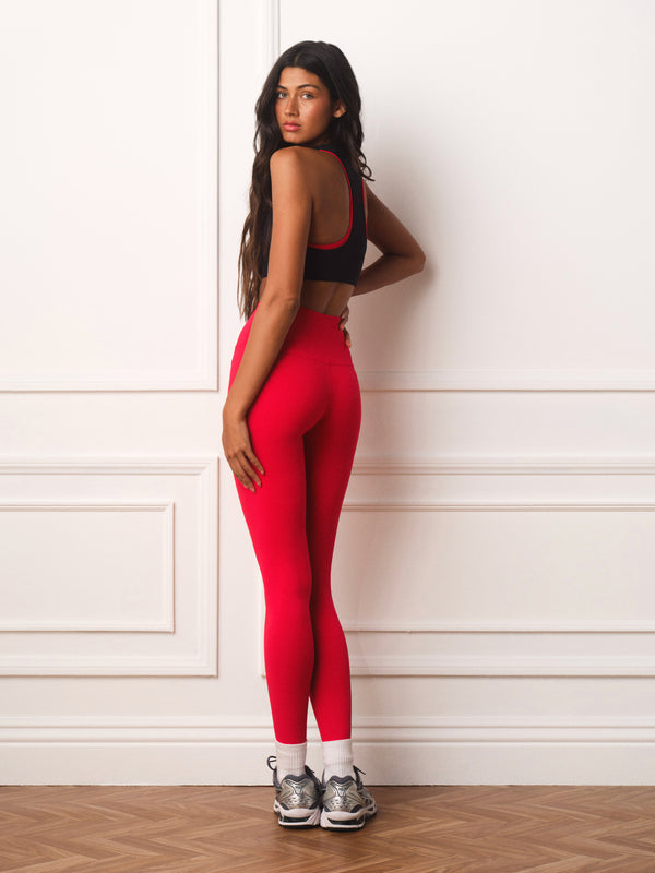 A woman stands in profile against a white paneled wall wearing bright red high-waisted leggings, a black sports bra with red trim, white socks, and gray athletic sneakers.