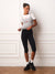 A woman modeling a light gray ribbed tank top with a small red logo, worn over a white t-shirt, paired with black capri leggings featuring white side stripes, white socks, and black sneakers, standing against a white paneled wall. <model-info>Courtney wears a size S and is 5’8”, 25” waist, 32B bust.</model-info>