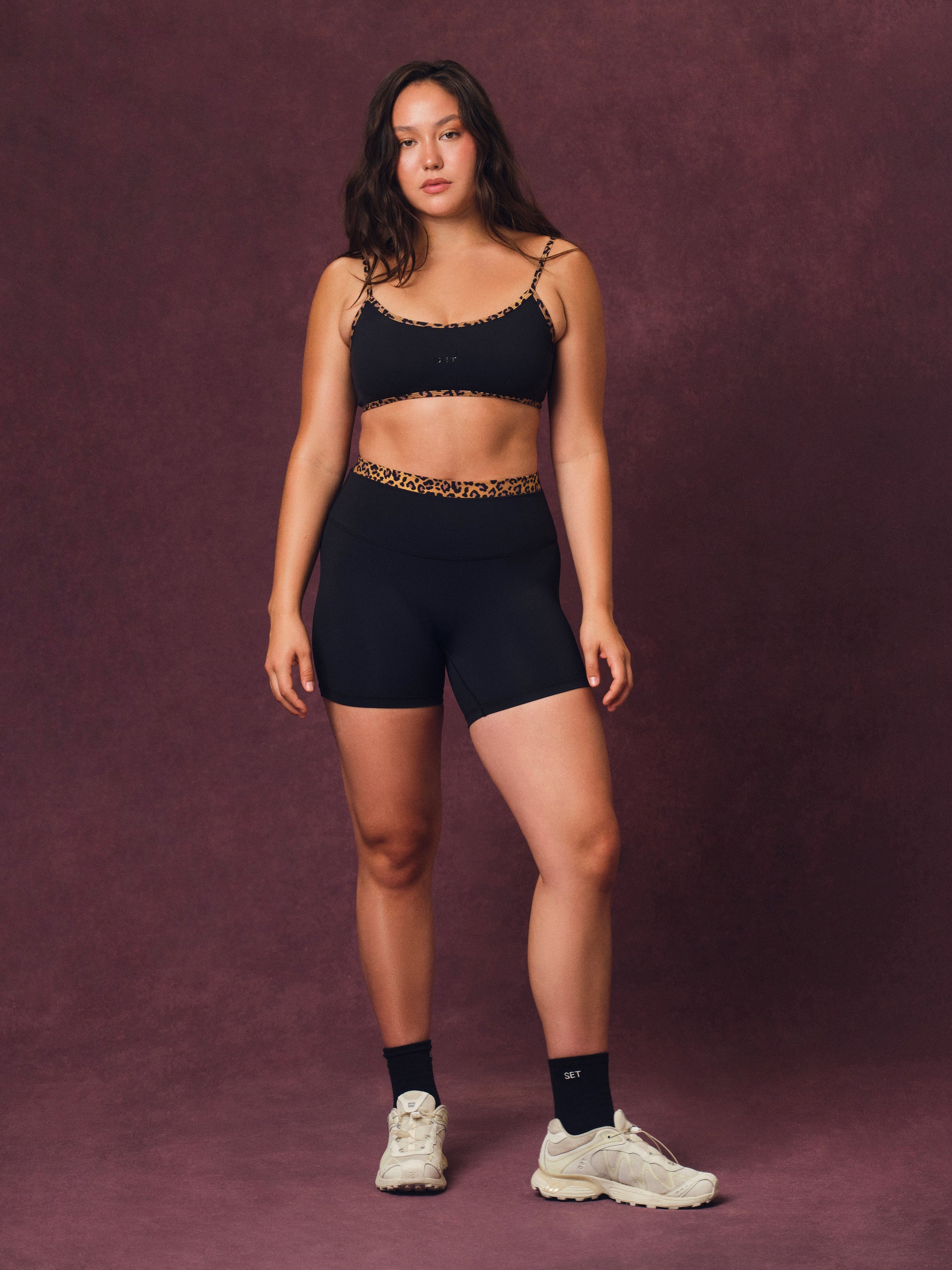 A woman modeling a black two-piece activewear set consisting of a cropped sports bra with gold leopard-print trim and matching high-waisted biker shorts, paired with black socks and white sneakers, against a textured dark purple studio backdrop.