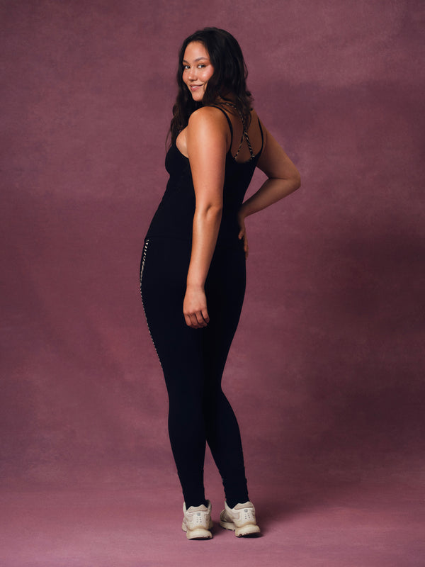 A woman modeling a black sleeveless jumpsuit with zebra detailing, standing against a textured mauve studio backdrop.