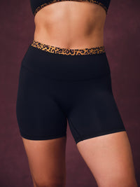 Black athletic biker shorts with a leopard print waistband, shown on a model from the back against a dark plum background. 
