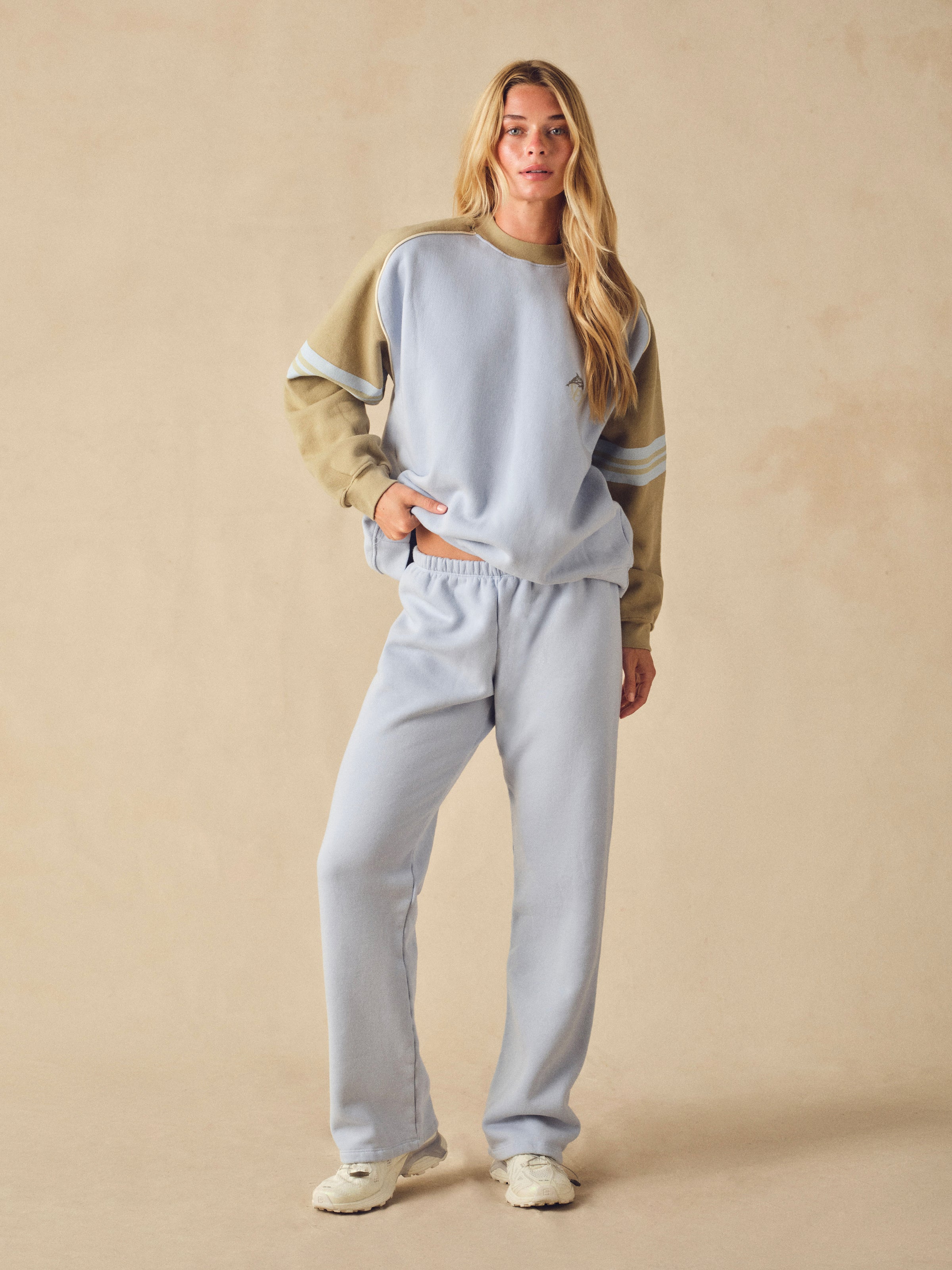 A woman modeling a light blue sweatshirt and matching sweatpants set with beige sleeves and shoulder details, standing against a neutral beige studio backdrop. 