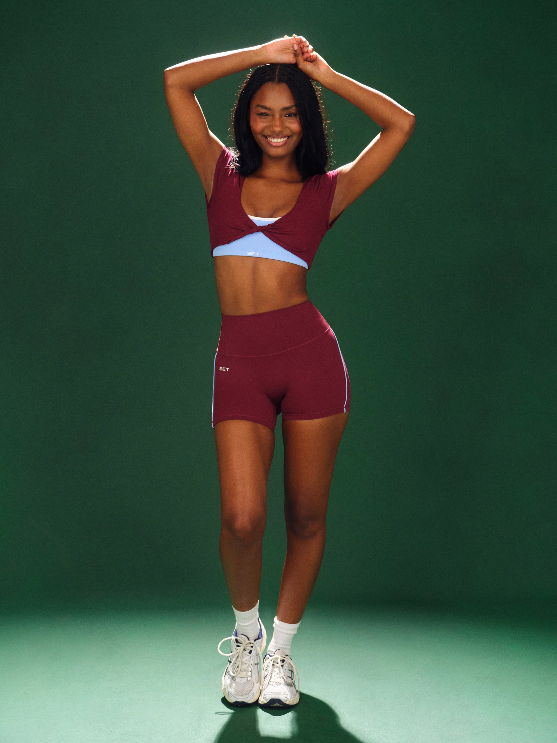 Woman in maroon athletic top and shorts posing against a green background