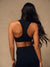 A woman shown from behind wearing a black sports bra and black leggings against a plain beige studio background.