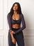 A woman modeling a dark grey long-sleeve crop top with a twist-front detail and black athletic bottoms, standing against a white paneled wall. <model-info>Meeya wears a size XS and is 5’8”, 23” waist, 32B bust.</model-info>