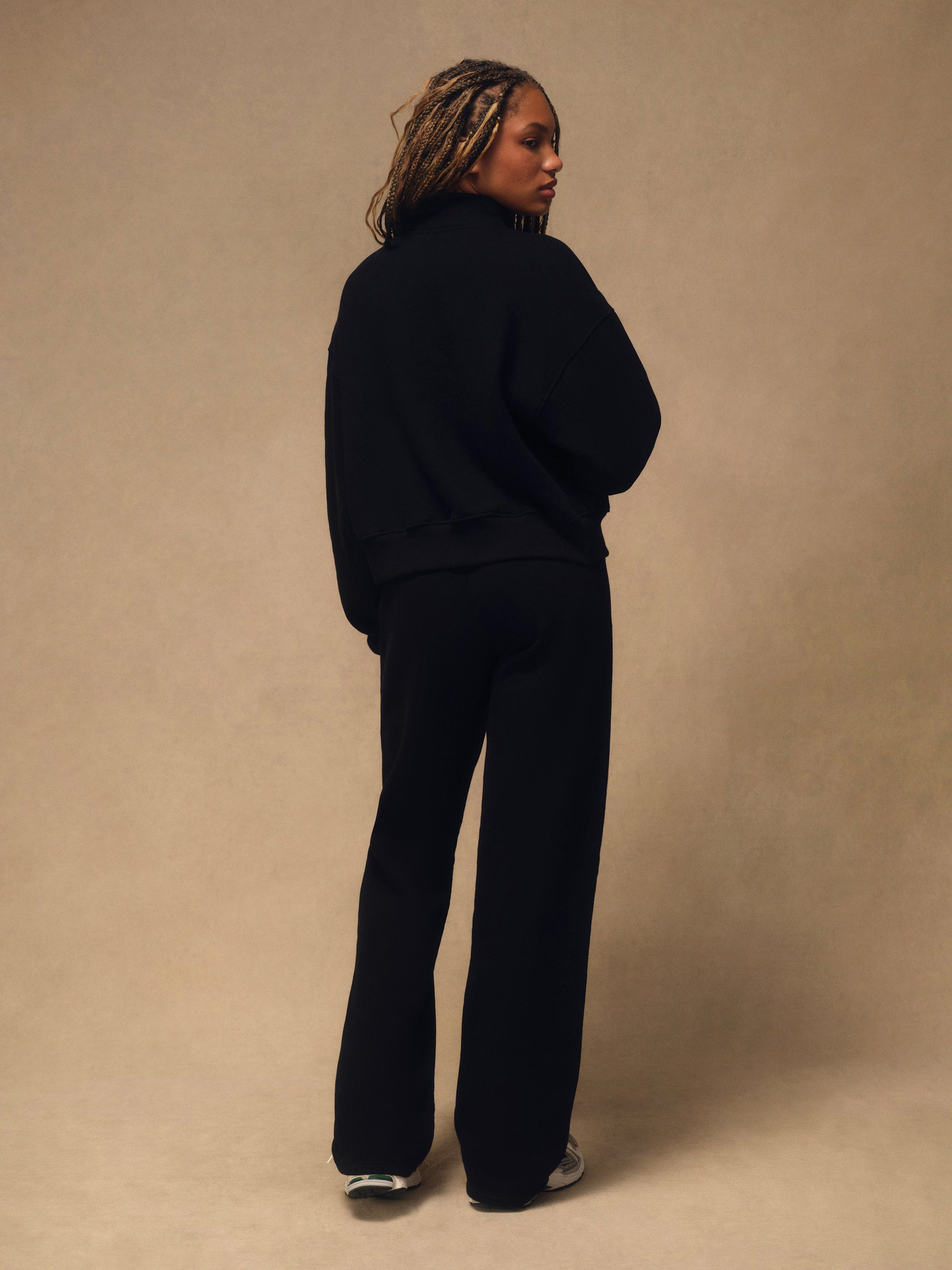 A model wearing a black sweatshirt and matching wide-leg trousers, photographed from the back against a warm beige studio backdrop.