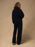 A model wearing a black sweatshirt and matching wide-leg trousers, photographed from the back against a warm beige studio backdrop.