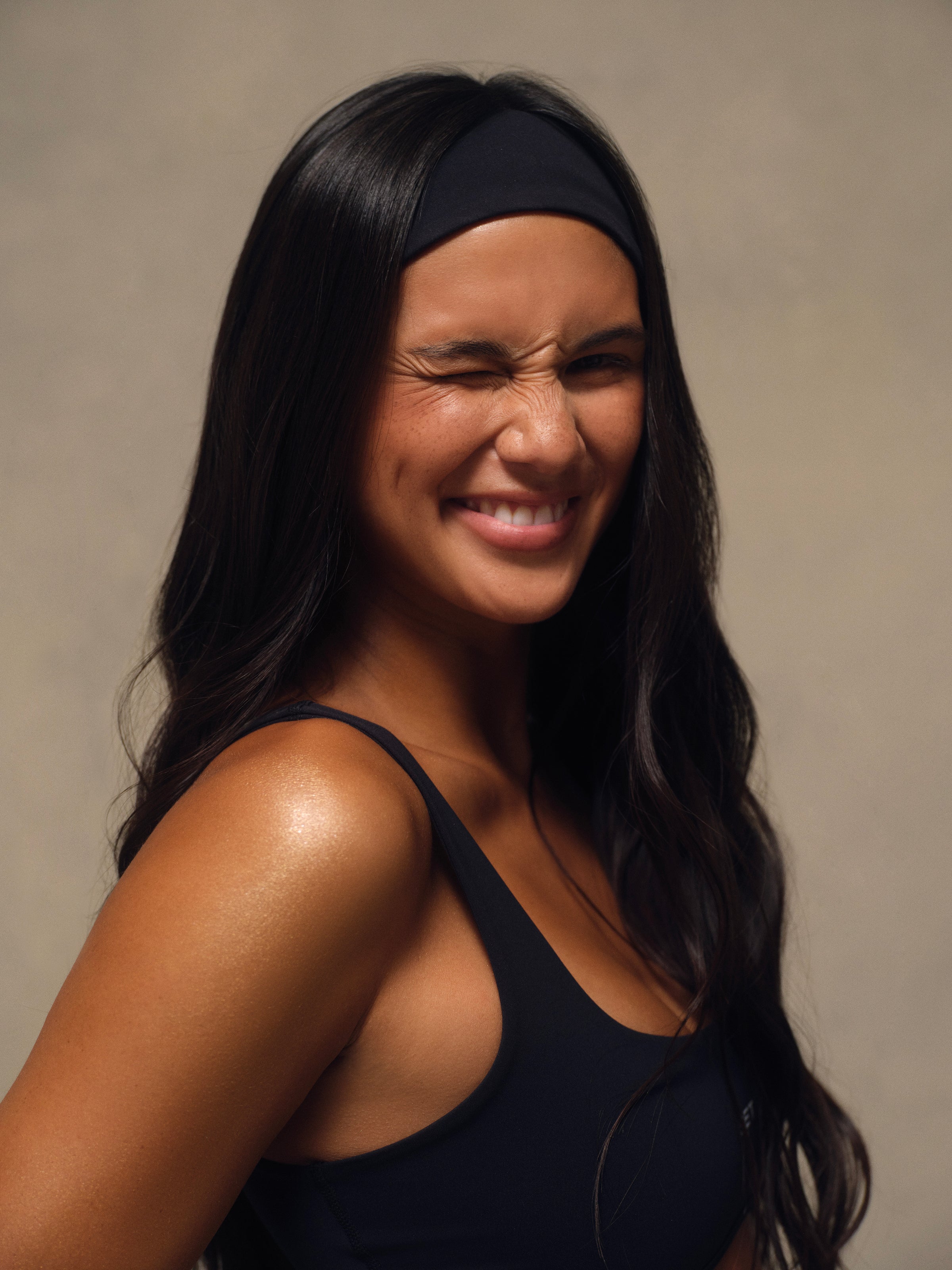 A woman with long dark hair wearing a black athletic headband and a black sports tank top, smiling and squinting against a neutral beige background.
