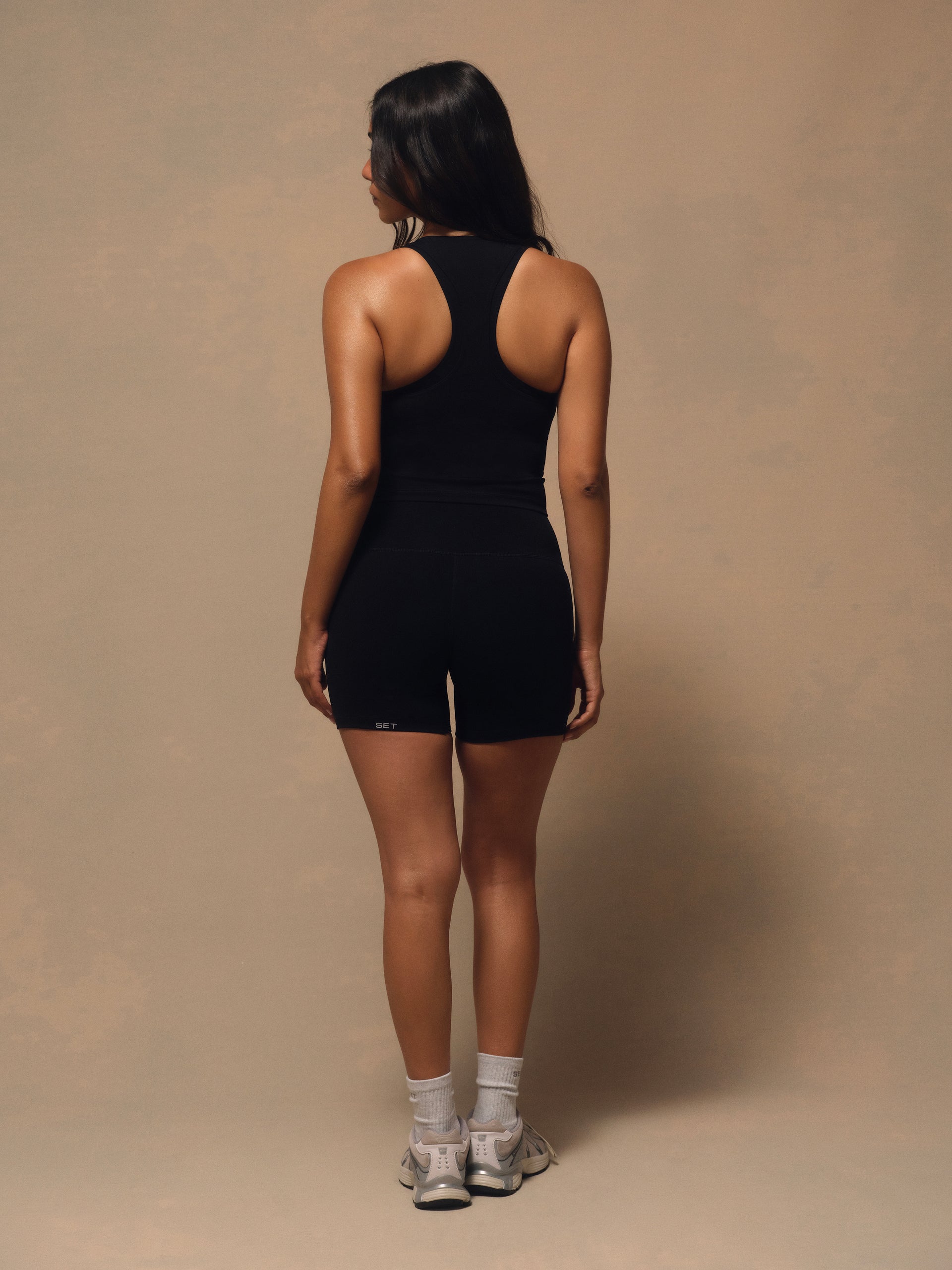 A woman is shown from behind wearing a black athletic tank top and matching black shorts, standing against a plain beige studio backdrop.