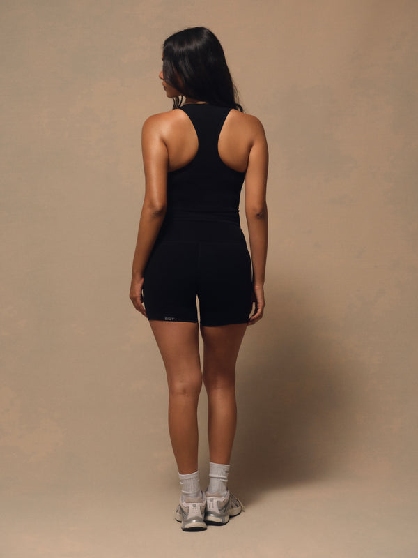 A woman is shown from behind wearing a black athletic tank top and matching black shorts, standing against a plain beige studio backdrop.