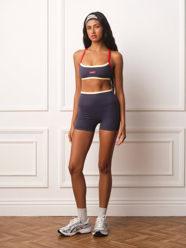 A woman modeling a matching athletic set consisting of a navy blue sports bra and biker shorts with red and beige trim, paired with white socks and gray sneakers, standing against a white paneled wall.