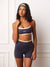 A woman modeling a matching athletic set consisting of a navy blue sports bra with cream and red trim, and matching high-waisted navy blue shorts with a cream waistband, against a white paneled wall. <model-info>Meeya wears a size XS and is 5’8”, 23” waist, 32B bust.</model-info>