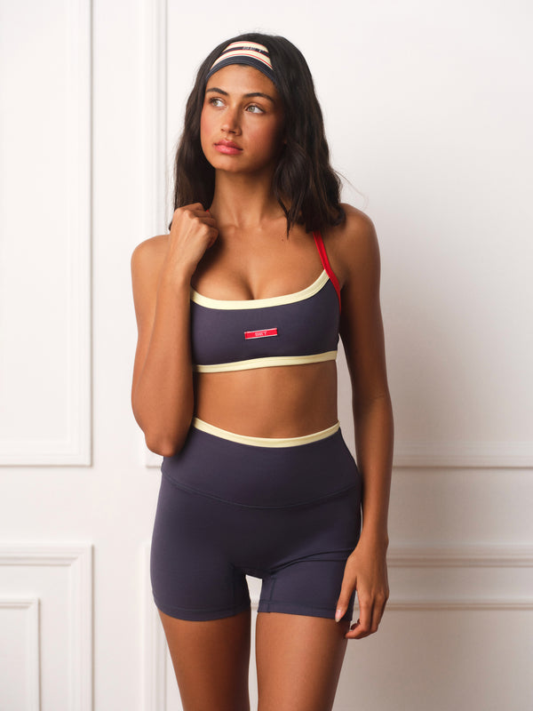 A woman modeling a matching athletic set consisting of a navy blue sports bra with cream and red trim, and matching high-waisted navy blue shorts with a cream waistband, against a white paneled wall.