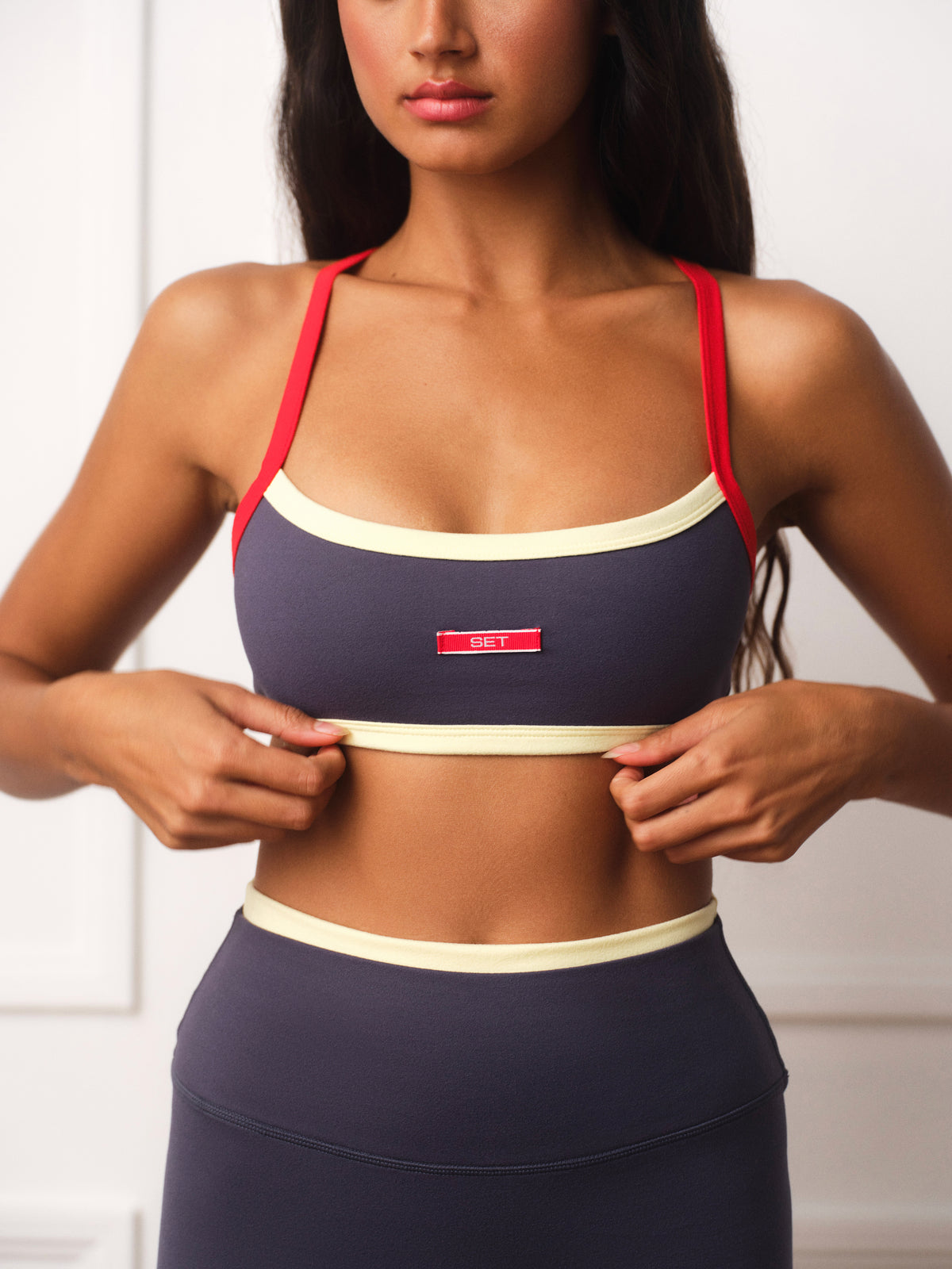 A woman wearing a navy blue sports bra with red straps and cream trim, holding the band down with both hands. The brand name 'SET' is visible on a small red label on the front.