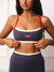 A woman wearing a navy blue sports bra with red straps and cream trim, holding the band down with both hands. The brand name 'SET' is visible on a small red label on the front.