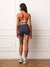 A woman stands with her back to the camera wearing a dark navy athletic bra with red and beige straps, paired with matching navy high-waisted shorts, white socks, and sneakers, set against a white paneled wall and wooden floor.