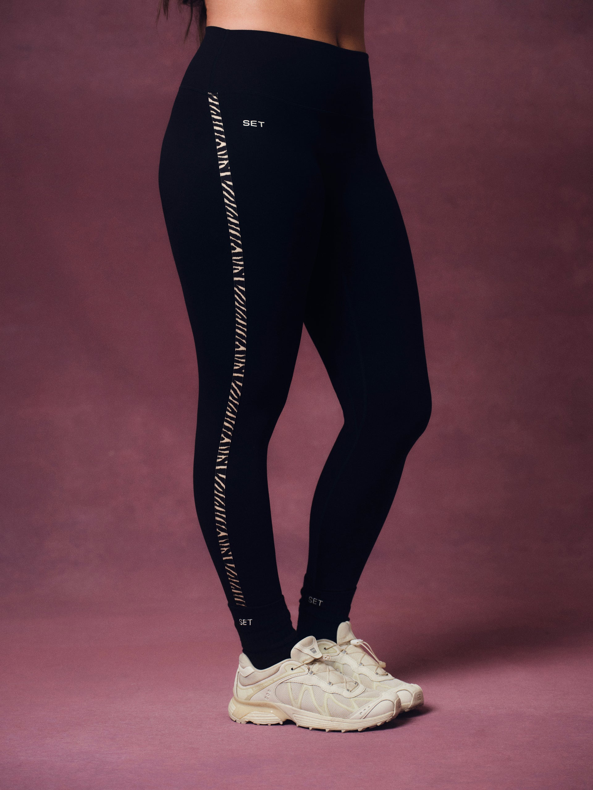 A person wearing black leggings with a vertical zebra print down the side, paired with off-white sneakers, standing against a solid mauve-pink studio backdrop.