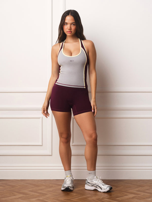 A woman modeling a light gray athletic tank top with dark side panels and matching dark burgundy athletic shorts, standing against a white paneled wall on a wood floor.