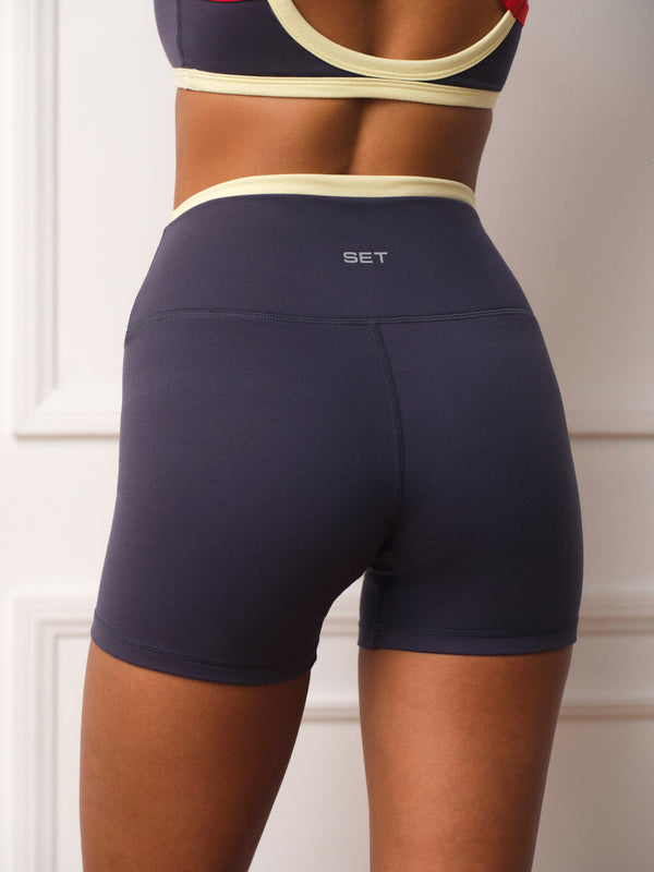 Rear view of a person wearing high-waisted navy athletic shorts with a cream waistband and a small white 'SET' logo, paired with a matching sports bra in a light interior setting.