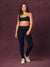 A woman modeling a black activewear set consisting of a sports bra and high-waisted leggings with cheetah patterned trim, paired with white sneakers, against a textured dark purple studio backdrop. <model-info> Christina wears a size L and is 5’9, 29½” waist, 36" bust.</model-info> 