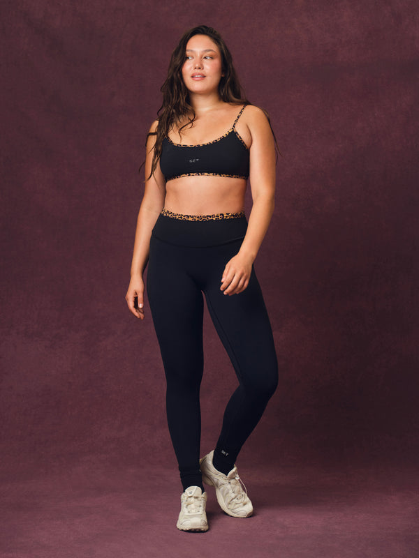 A woman modeling a black activewear set consisting of a sports bra and high-waisted leggings with cheetah patterned trim, paired with white sneakers, against a textured dark purple studio backdrop. 