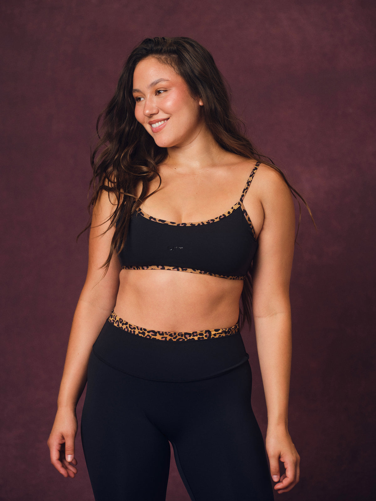 A woman modeling a matching black activewear set with leopard print trim, consisting of a sports bra and high-waisted leggings, against a textured dark purple studio backdrop. <model-info> Christina wears a size L and is 5’9, 29½” waist, 36" bust.</model-info> 