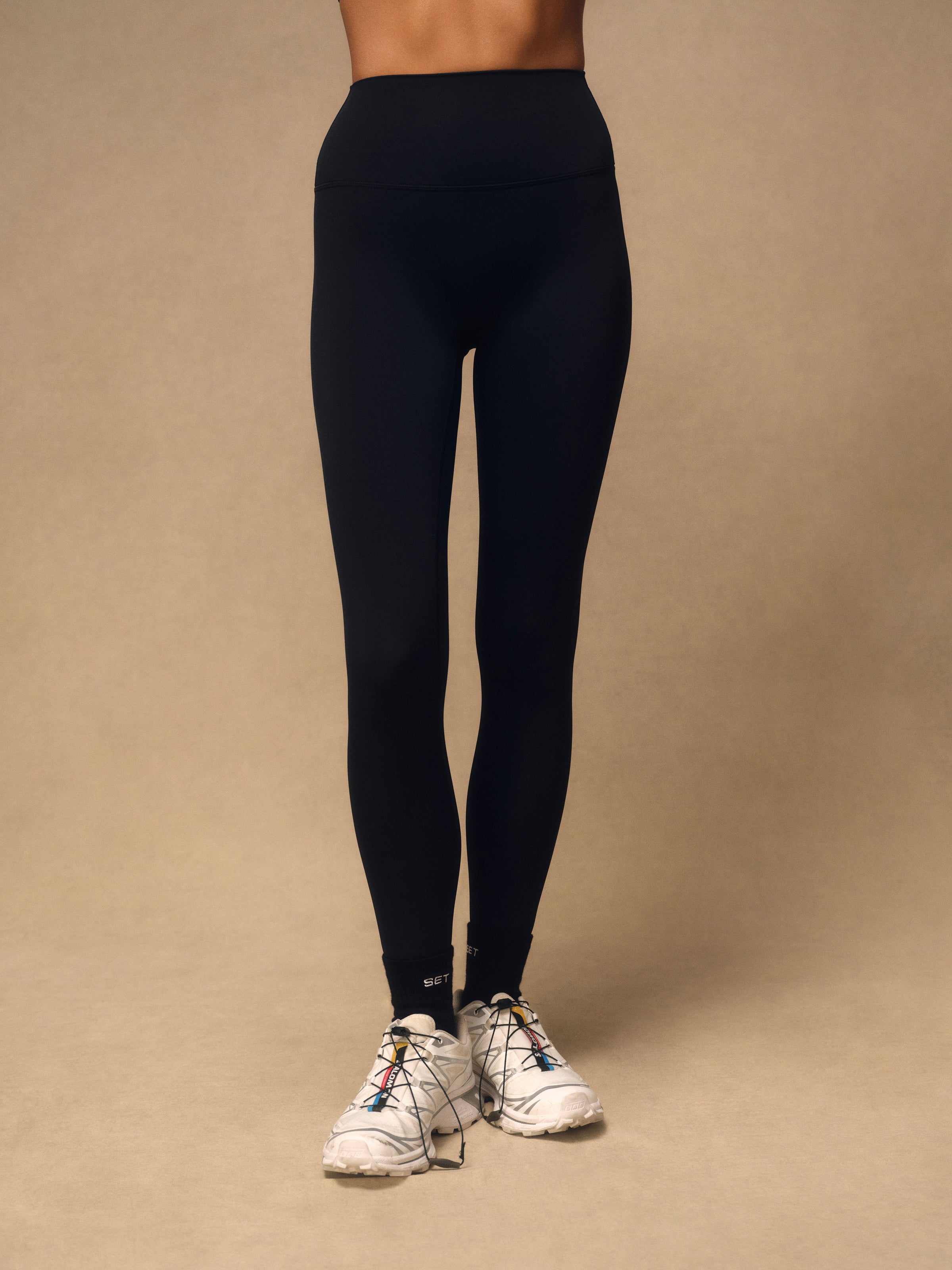 A model from the waist down wearing black high-waisted leggings and white trail running shoes, standing against a solid beige studio background.