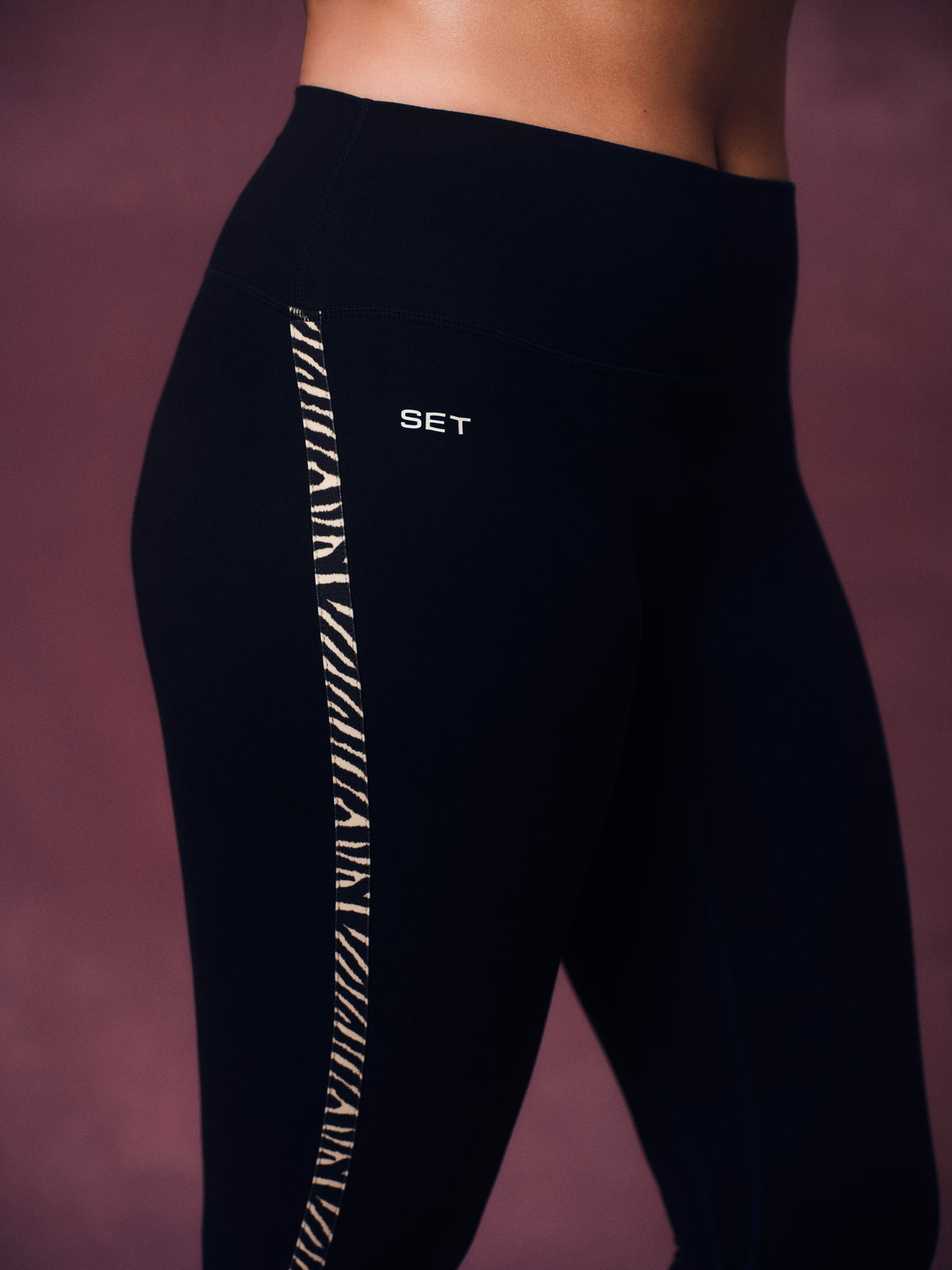 Close-up of a model wearing black high-waisted leggings with a vertical white stripe featuring zebra pattern and a small white 'SET' logo on the hip, against a dark mauve studio background.