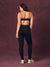 A woman shown from the back wearing a black sports bra with gold patterned straps and a matching black high-waisted leggings set, standing against a textured dark purple studio backdrop.