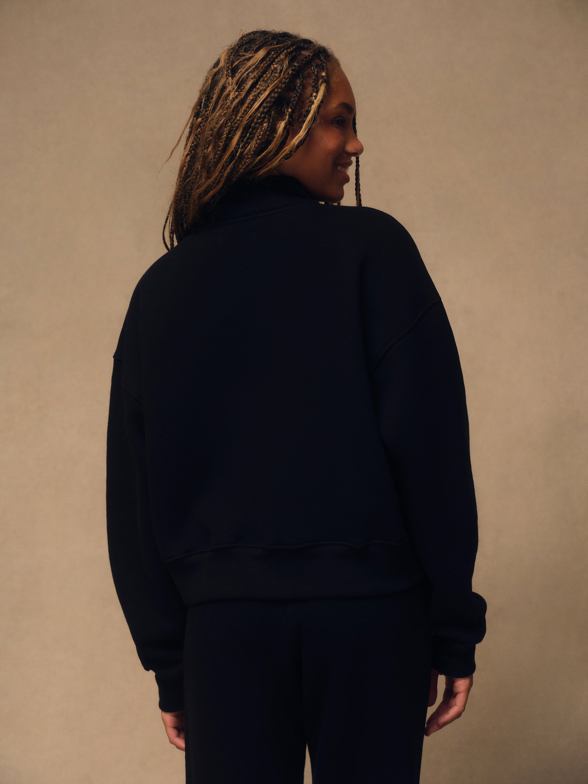 A woman with blonde braids is shown from behind wearing a black crewneck sweatshirt and matching black pants against a neutral beige background.