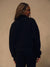 A woman with blonde braids is shown from behind wearing a black crewneck sweatshirt and matching black pants against a neutral beige background.
