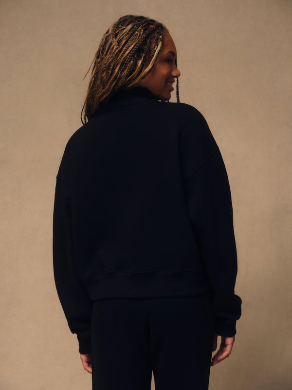 A woman with blonde braids is shown from behind wearing a black crewneck sweatshirt and matching black pants against a neutral beige background.