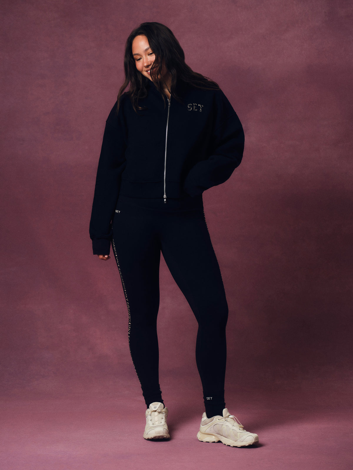 A woman modeling a matching black zip-up hoodie and leggings set with white sneakers, standing against a textured mauve-pink studio backdrop.