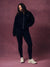 A woman modeling a matching black zip-up hoodie and leggings set with white sneakers, standing against a textured mauve-pink studio backdrop.