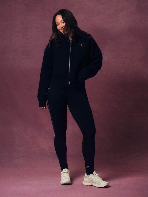 A woman modeling a matching black zip-up hoodie and leggings set with white sneakers, standing against a textured mauve-pink studio backdrop.