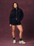 A woman modeling a black zip-up hoodie and matching black shorts with small gold 'SET' branding, styled with black socks and off-white sneakers against a textured dark mauve studio  <model-info> Christina wears a size L and is 5’9, 29½” waist, 36" bust.</model-info> backdrop.