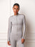 A woman modeling a light gray long-sleeve athletic zip-up jacket and matching leggings. The jacket features dark red and cream stripes down the sleeves and a small 'WEST' logo on the chest, set against a white paneled wall background. <model-info>Courtney wears a size XS and is 5’8”, 25” waist, 32B bust.</model-info>