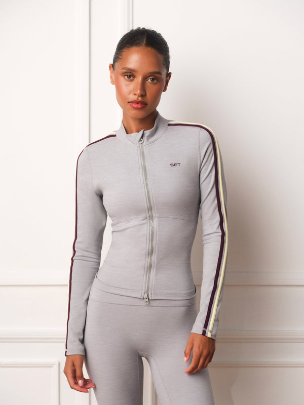 A woman modeling a light gray long-sleeve athletic zip-up jacket and matching leggings. The jacket features dark red and cream stripes down the sleeves and a small 'WEST' logo on the chest, set against a white paneled wall background.