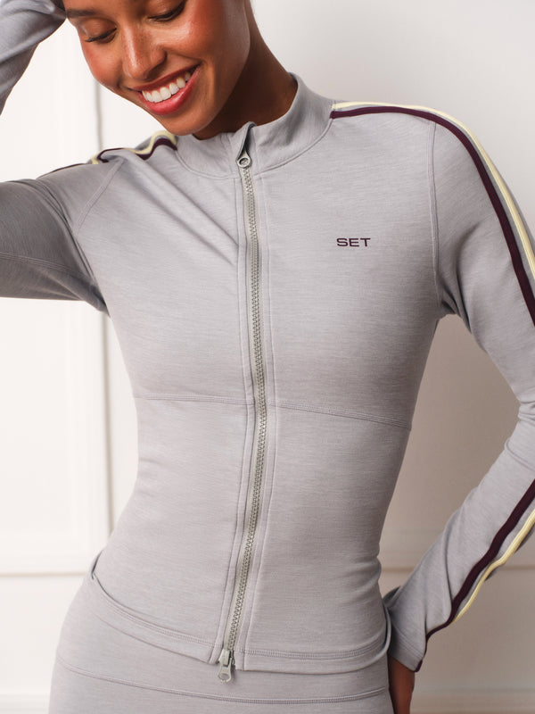 A woman wearing a light gray long-sleeve athletic zip-up jacket with dark burgundy and cream stripes down the sleeves and a small 'SET' logo on the chest, paired with matching gray bottoms.